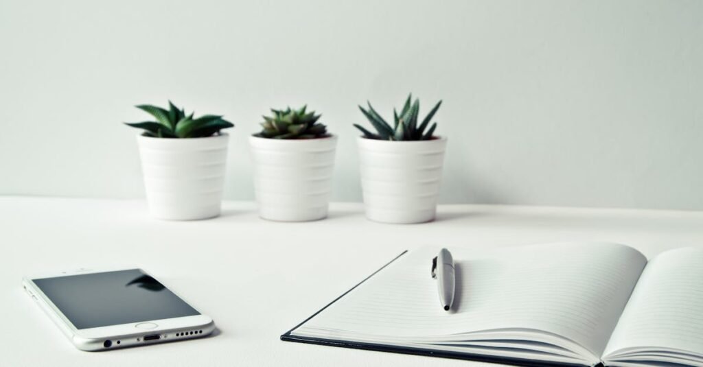 A clean, minimalist workspace featuring a smartphone, open notepad, pen, and potted succulents.